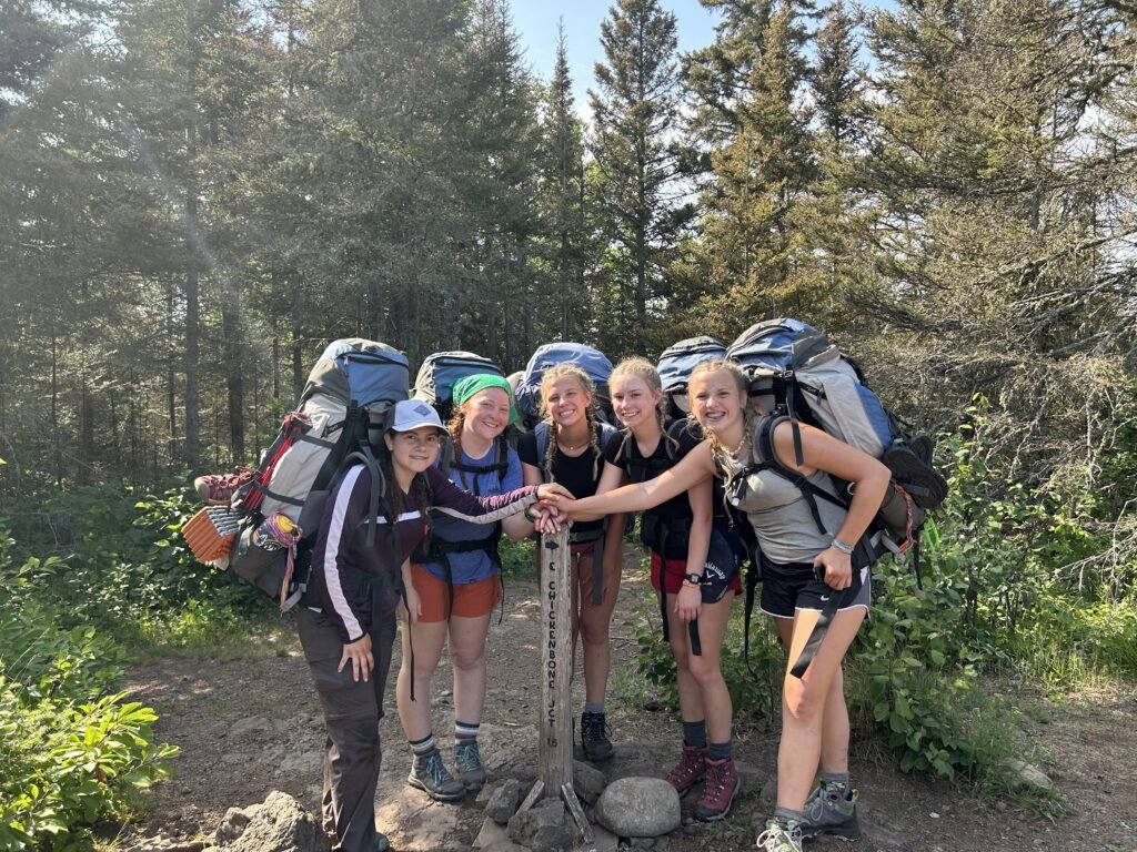 Photo of five smiling young women wearing backpacks with their hands piled on a trail junction sign in the woods.