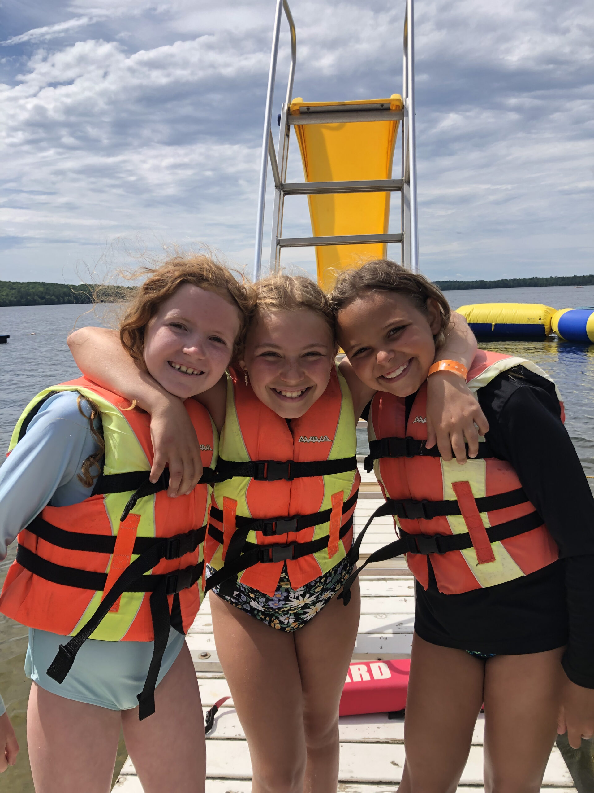 Three young girls smile brightly for a photo on a white dock at a lake. They are huddled close together with their arms around each other's shoulders, all wearing matching orange and yellow life jackets.