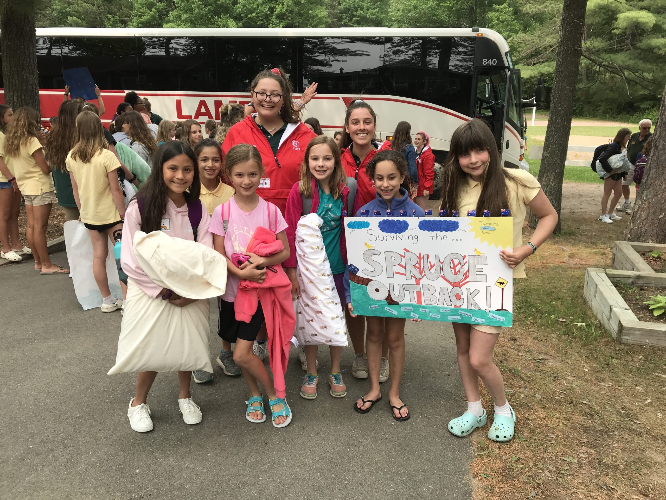 A group of smiling young campers and two staff members pose for a photo in front of a large white and red bus.
