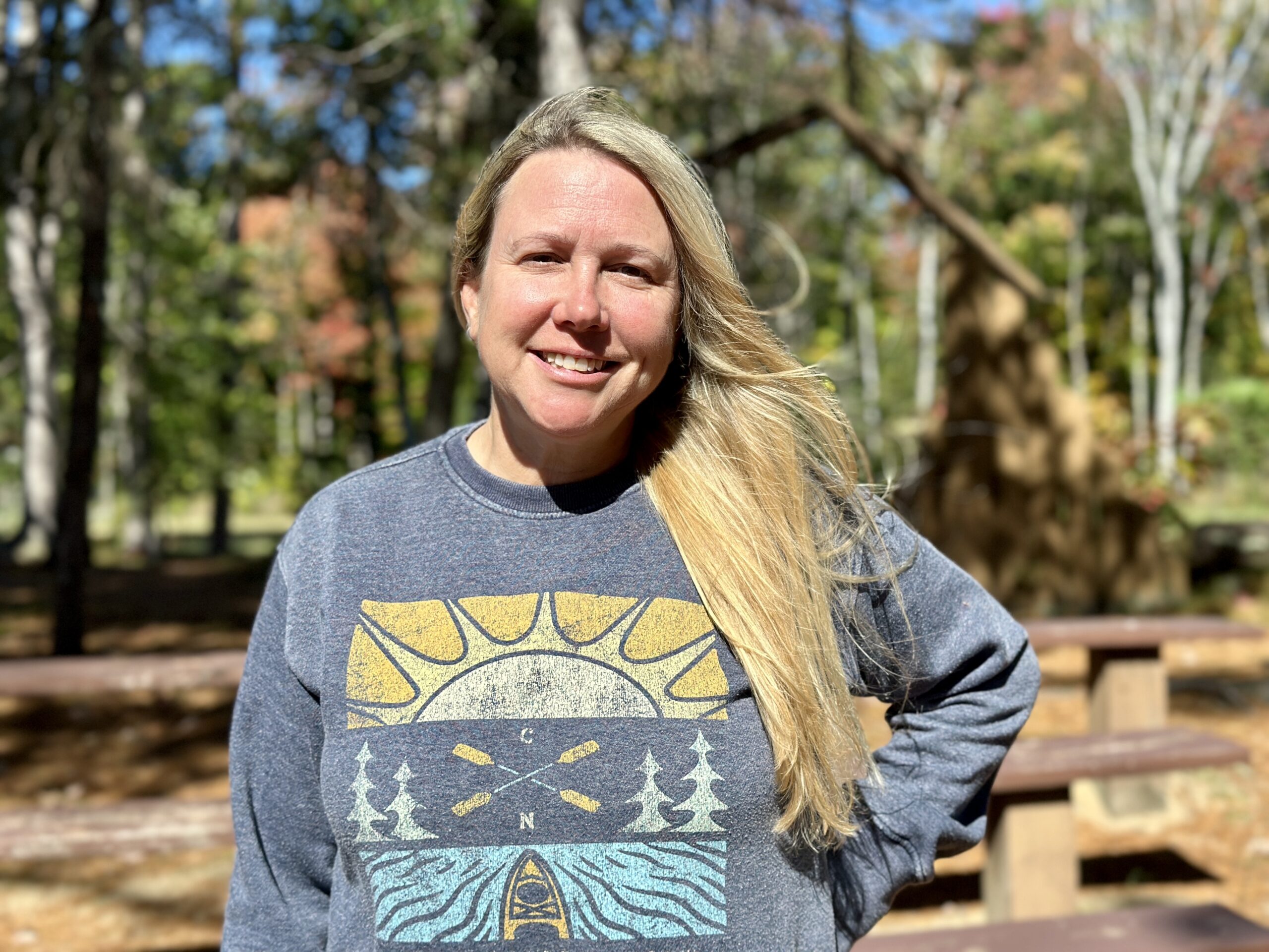 A smiling woman with long blonde hair, Camp Nicolet co-director Laura, stands outdoors in a sunny, wooded area.