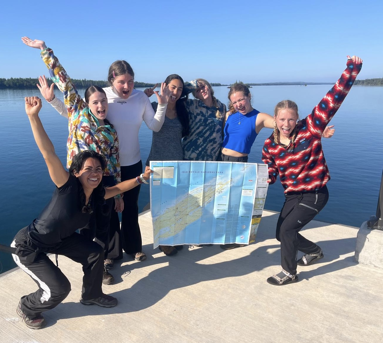 Gemini said A group of seven energetic young women pose excitedly on a concrete pier overlooking a vast, calm blue lake under a clear sky. Two of them hold up a large, unfolded map of Isle Royale National Park.
